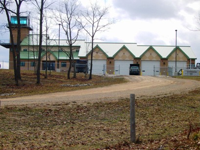 This picture of the Range 40 Support Complex was taken from the entrance road, through winter-bare trees.  The building has light and dark tan masonry walls with green metal roof and trim.  The blue tint of the control tower window provides contrast against the grey winter sky.