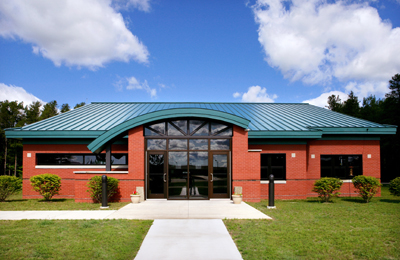 picture of Billeting Services Building at Alpena Combat Readiness Training Center - red brick walls with cream colored masonry accents, blue/green metal roof, tinted windows and curved metal 'eyebrow' detail over main entrance doors