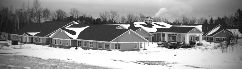exterior photo of Mill Creek Assisted Living Facility in Marquette MI - this panoramic black & white winter picture shows much of the facility surrounded by snow-covered land against a backdrop of winter bare trees