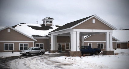 exterior photo of Mill Creek Assisted Living Facility in Marquette, Michigan - this Winter picture is of the front entry portico - the facility has taupe colored siding, white columns and trim, and a dark brown and grey architectural shingled roof