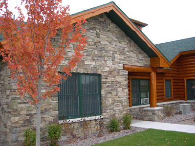 exterior photo along front elevation of Freighter View Assisted Living Facility in Sault Ste. Marie, Michigan - mixed stone siding, natural log siding with timber frame elements, limestone sills and ledges, with green shingled roof and coordinating green eaves and window trim