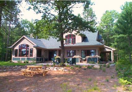 picture of waterfront custom home - this exterior photo is taken on the lake side elevation of the home - light wood siding, green architectural shingle roof, dark red window trim and plenty of outdoor living area