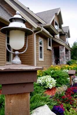 picture of lakeside custom home on Otsego Lake - this is an exterior photo taken at an angle along the front of the home, with fence-post lantern in the foreground - home has natural wood siding, architectural shingled roof and fieldstone columns at porch