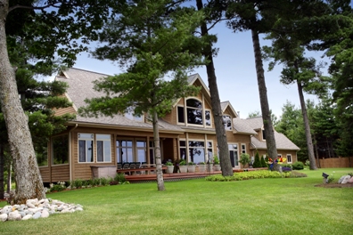 picture of lakeside custom home on Otsego Lake - this exterior photo was taken of the lakeside elevation - custom windows with unique trim, multiple sets of french doors leading onto outdoor decks shaded by mature evergreens overlooking a sweeping lawn leading down to the water's edge