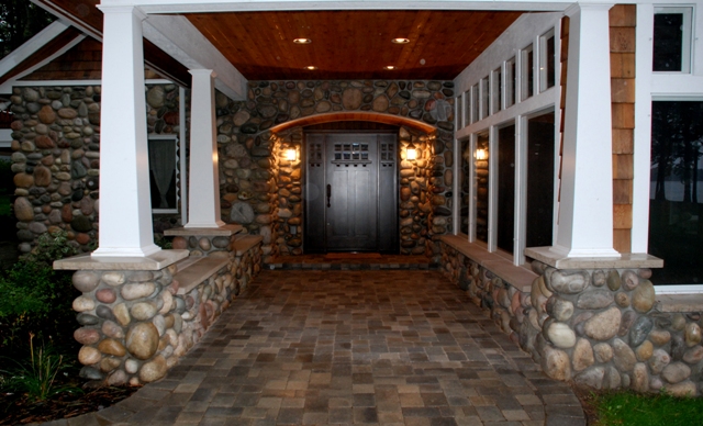 exterior photo of main entry to lakeside home in Lewiston, Michigan - covered porch with white columns against fieldstone exterior, wood plank ceiling with drop lights and wall sconces flanking custom entry door