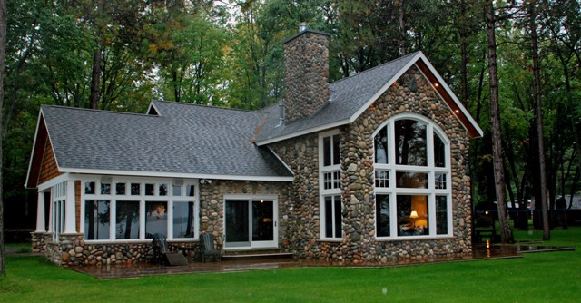 exterior photo of lakeside elevation of fieldstone home with white window and door trim, and grey architectural shingle roof