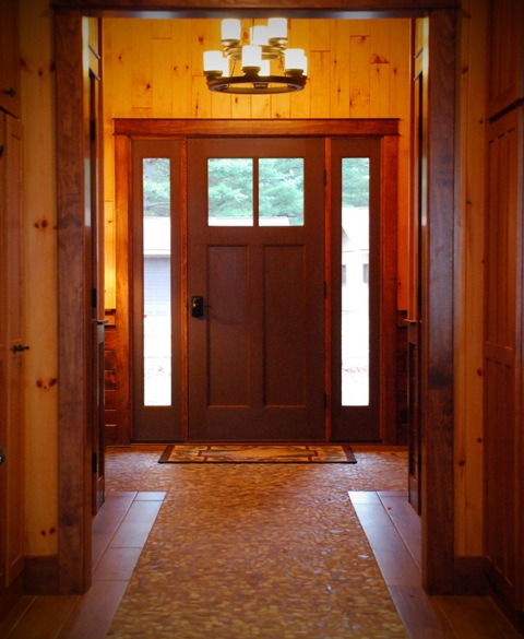 interior photo looking toward front entry, light wood plank walls, dark wood trim and tiered pendant light fixture over pebble-stone mosaic floor