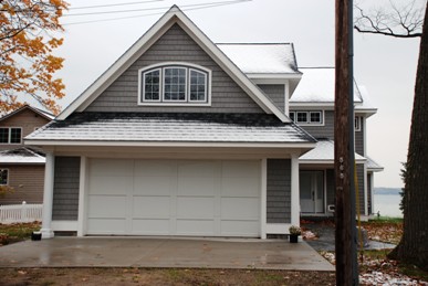 picture of lake side home in Topinabee, Michigan - this exterior photo facing the garage is taken from the road-side elevation, with the lake in the background - a light dusting of snow has fallen which complements the home's dark grey siding and roof, white garage doors and white trim