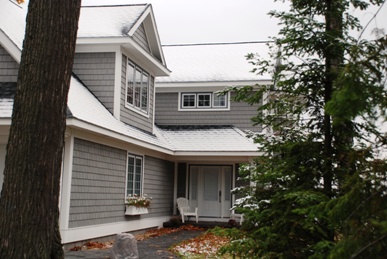 picture of lakeside home in Topinabee, Michigan - this exterior photo is looking through trees at the home's main entry showing snow-dusted roof, dark grey cedar siding and white trim, white wood-and-glass entry door and sidelights, with white rocking chair on porch