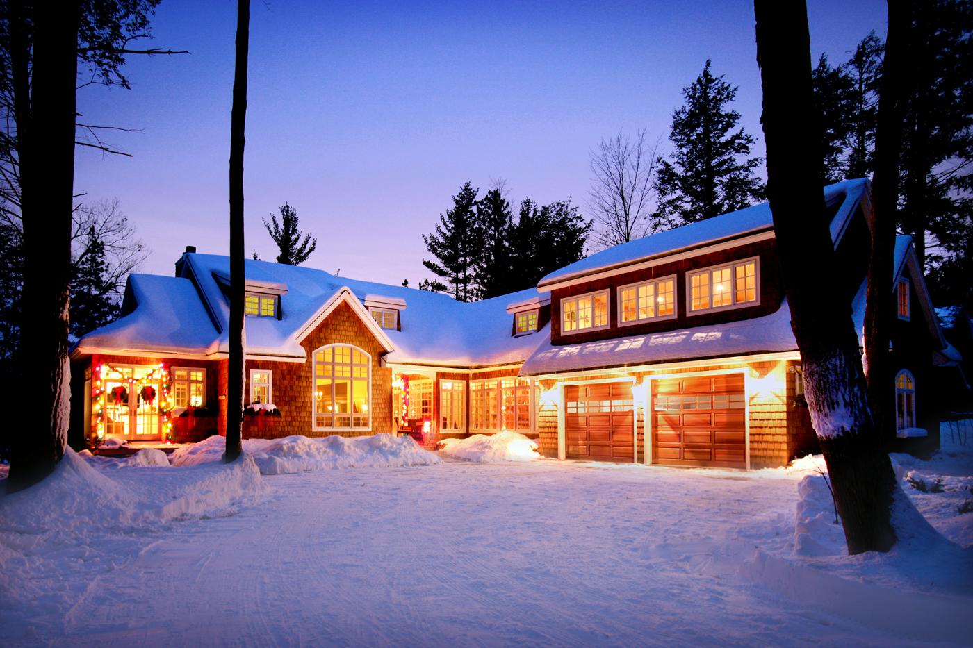 picture of lakeside custom home on O'Rourke Lake - this winter evening exterior photo shows the home's lights reflecting off the snow, showcasing the home's beauty - warm lighting illuminating the natural cedar siding, white trim and roof blanketed with snow