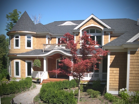 picture of custom home on West Bay, Traverse City, MI - this exterior photo shows the cottage's natural cedar siding with grey architectural shingle roof and white trim, curved window construction, curved arch over front entry porch which is repeated in a curved bump-out in the roof construction above